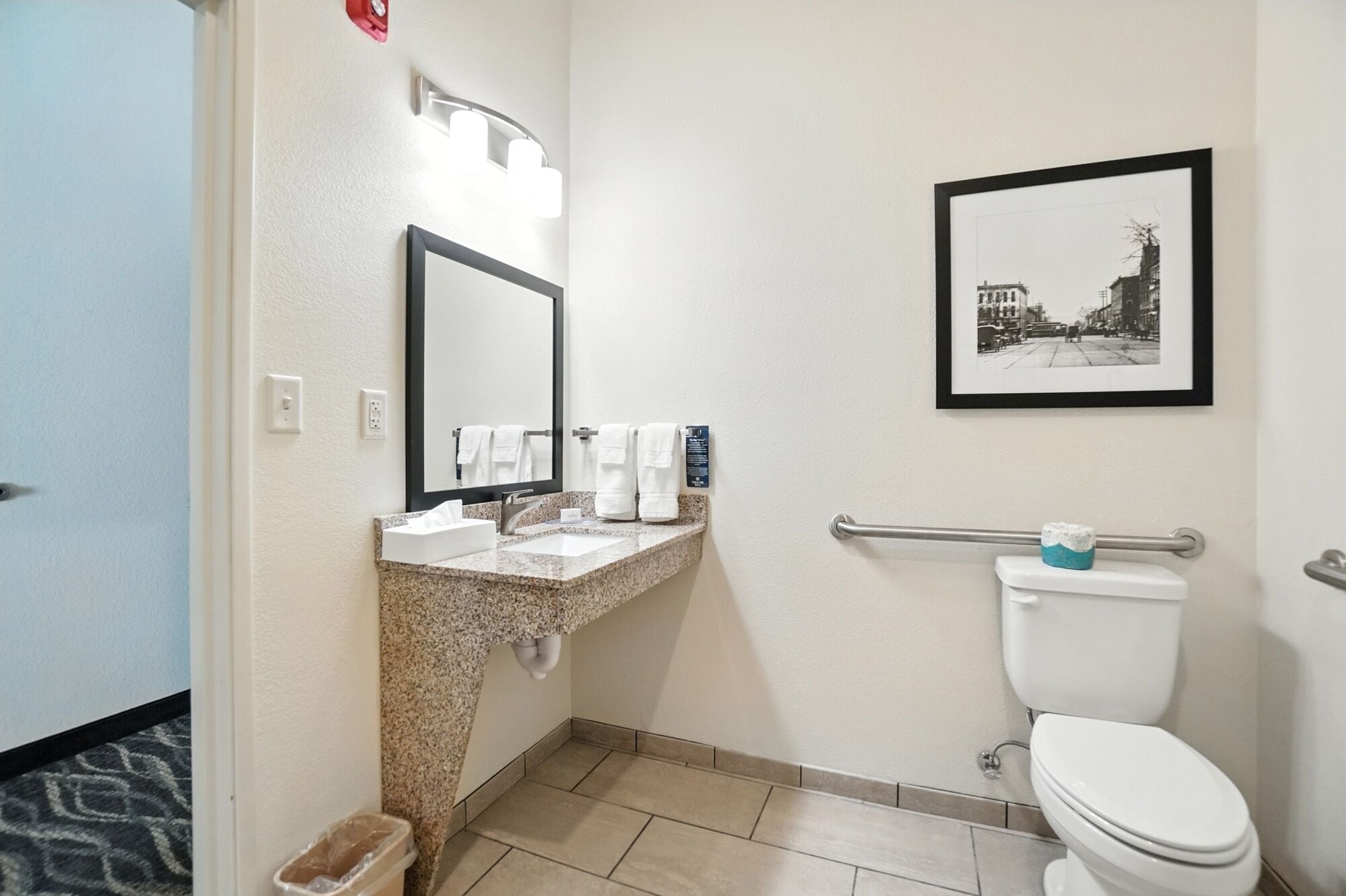Un baño pequeño con lavamanos de encimera de granito, inodoro blanco y un cuadro enmarcado con una vista urbana.
