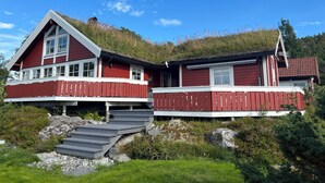 Una casa de madera roja con techo inclinado y balcón, rodeada de follaje y rocas.