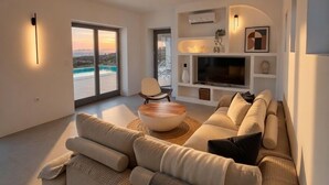 Un salón moderno con un sofá grande, una mesa de centro y un televisor. Se ve el exterior a través de amplios ventanales.