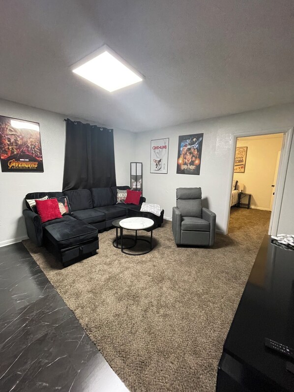 A living room with a sectional sofa, a grey armchair, a coffee table, and a flat-screen TV. There are posters on the walls and a ceiling light.