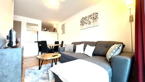 A modern living room with a grey sofa, a wooden coffee table, and a TV on a stand. There is a dining area with a table and chairs in the background. - Apartment for 6 people (Allos)