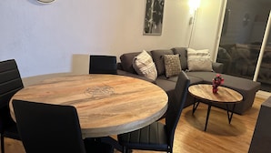 A modern living room with a round wooden table, black chairs, a grey sofa with patterned cushions, and a small side table with a decorative plant. - Apartment for 6 people (Allos)