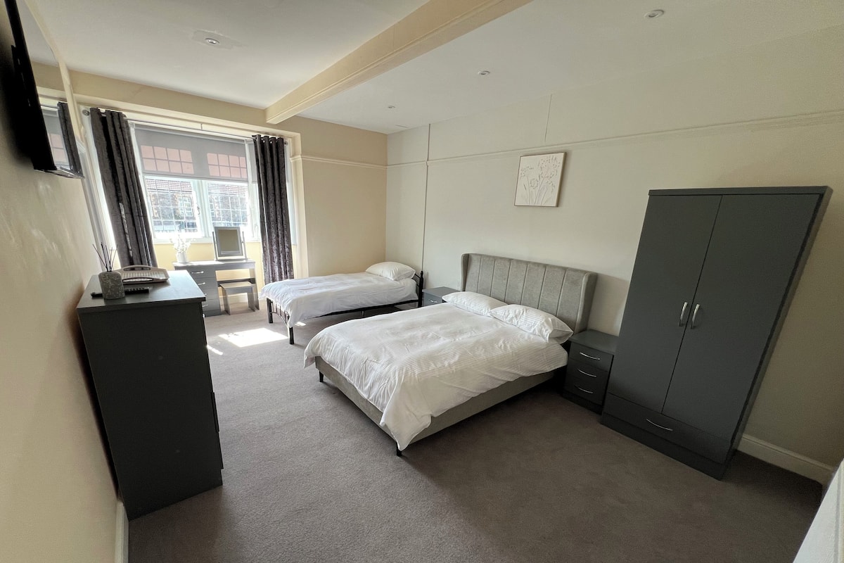 Ein Hotelzimmer mit zwei Einzelbetten, einem Schreibtisch mit Stuhl, einem Kleiderschrank und einem Fenster mit Vorhängen.