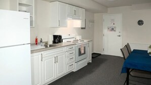 Una cocina compacta con armarios blancos, un refrigerador blanco, una estufa y un lavamanos. Hay una mesa azul con sillas y un reloj en la pared.