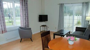 Un salon avec une table à manger en bois, un fauteuil gris, un canapé, un téléviseur fixé au mur et une fenêtre avec des rideaux.