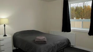 A bedroom with a bed, a nightstand, a lamp, a window with curtains, and a radiator.