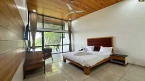 A bedroom with a wooden ceiling, a bed with white bedding, a nightstand, a desk with a chair, and a large window with curtains. - Bansari Greens Resort (Aluva)