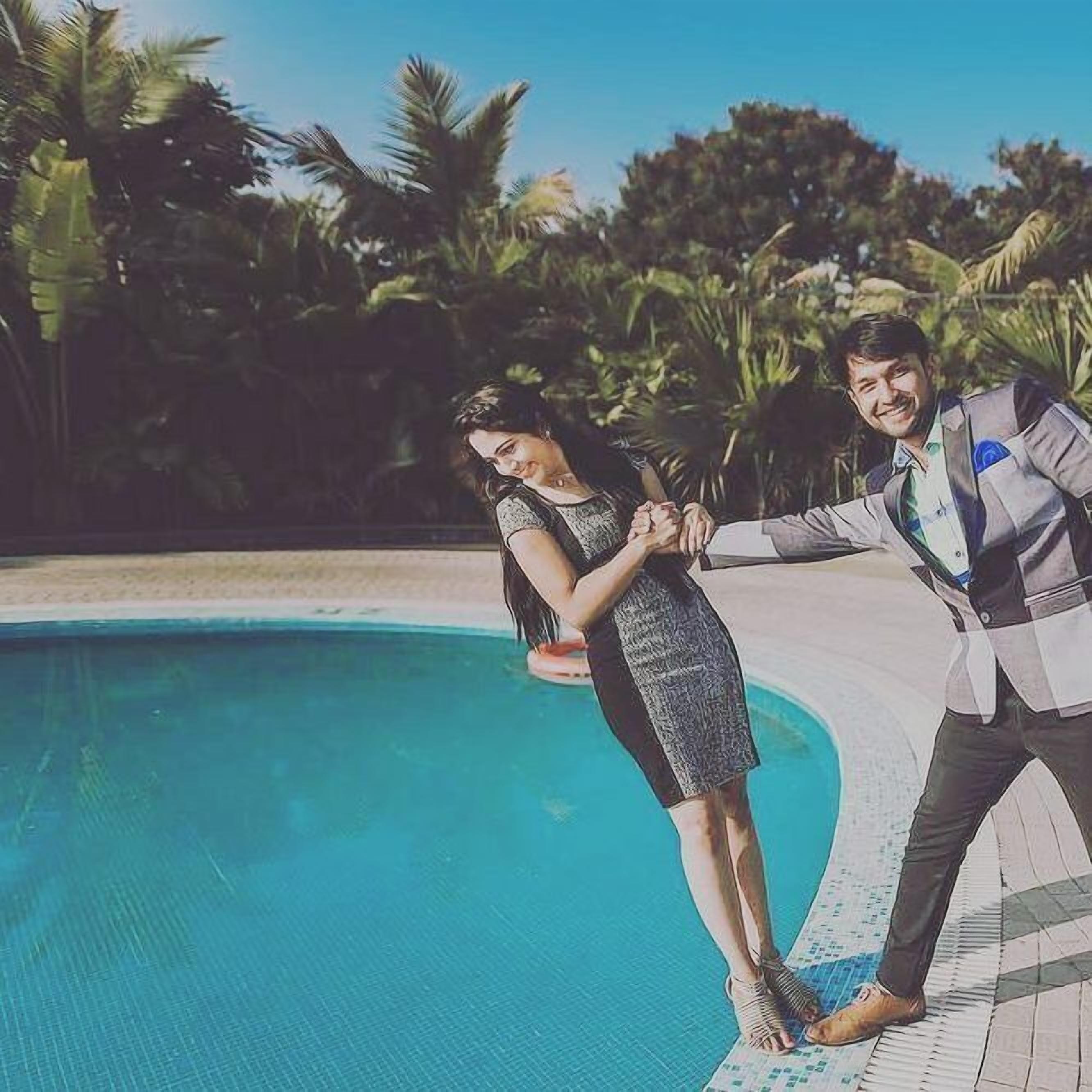 Una pareja parada junto a una piscina, con palmeras al fondo.
