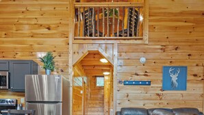 A wooden cabin interior with a kitchen, a dining area, a living room with leather sofas, and a loft bedroom.
