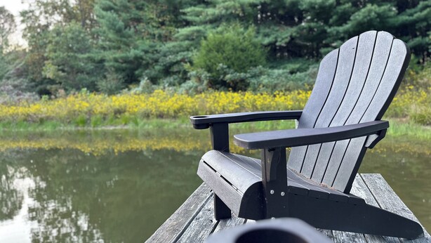 Een houten steiger met een zwarte Adirondack-stoel gericht op een waterpartij, omgeven door dichte groene bomen en gele bloemen.
