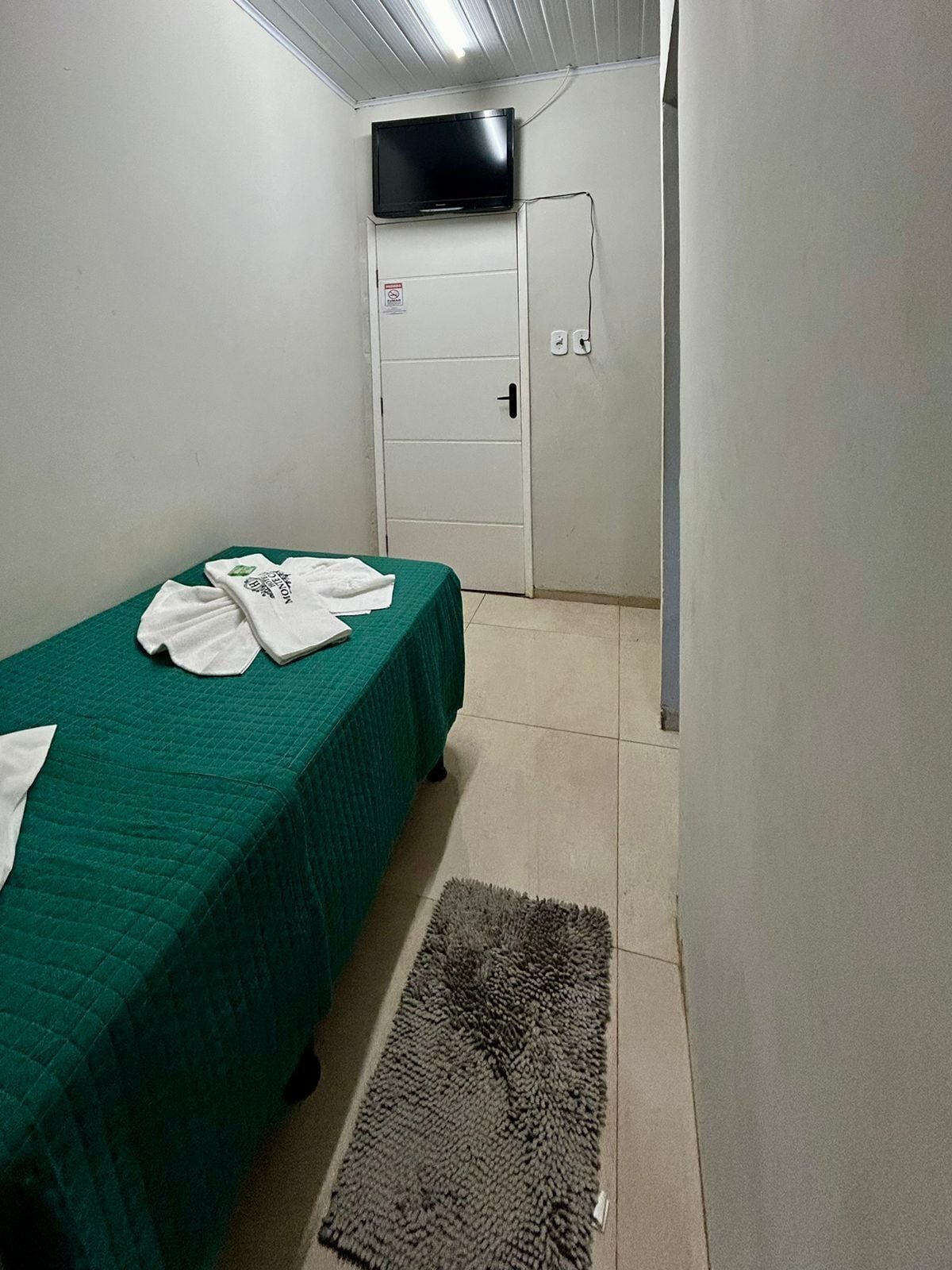 A small, clean room with a single bed, a TV mounted on the wall, and a door with a sign.