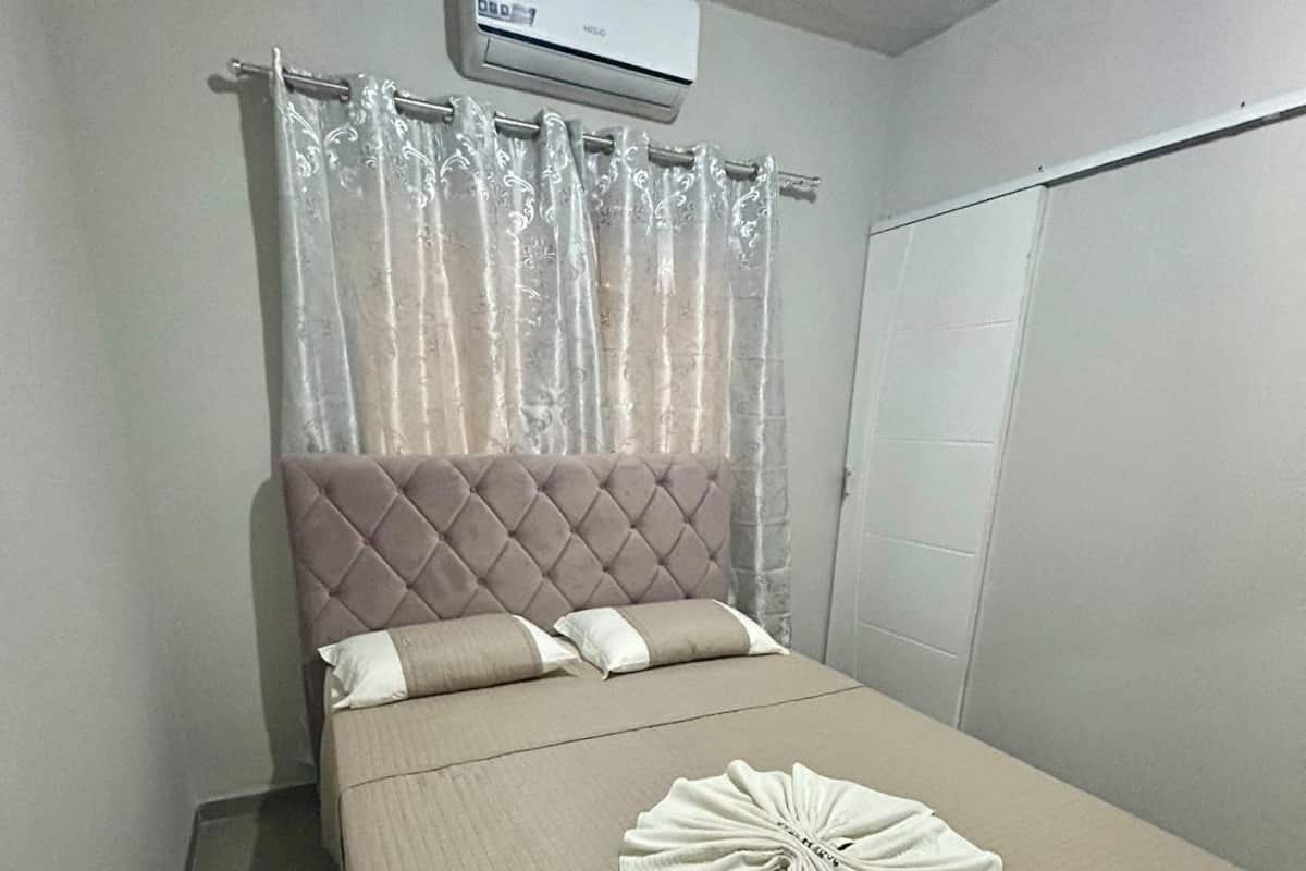 A neatly made bed with a quilted cover, a decorative white towel on the bed, a tufted headboard, and a white air conditioning unit above the bed.