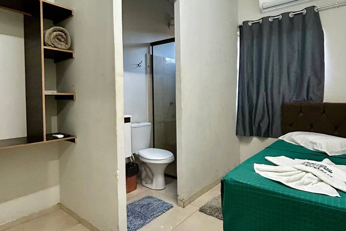A small, clean room with a bed, a toilet, and a shower. There are shelves with towels and a curtain over the shower.