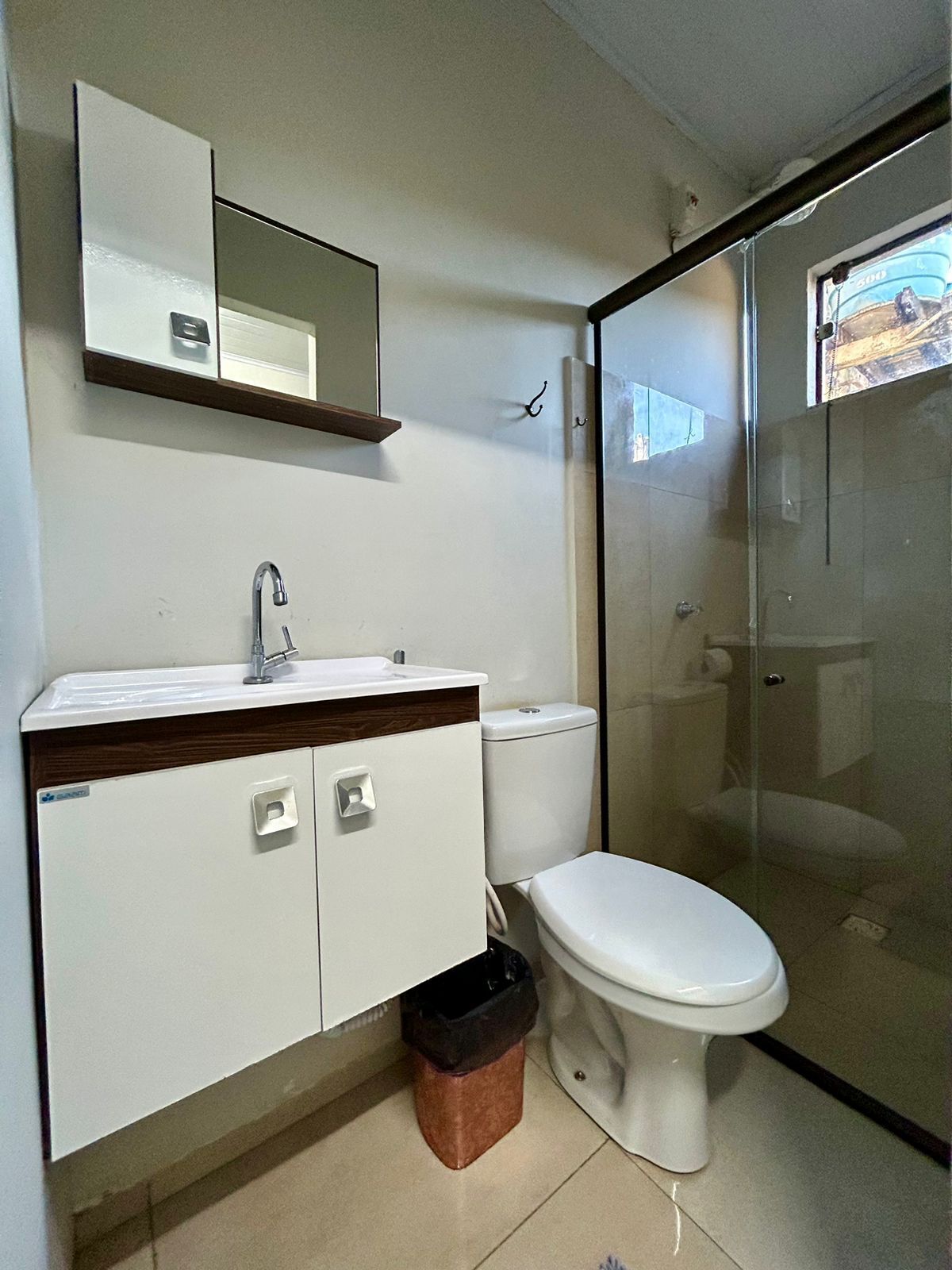 A bathroom with a white sink, mirror, and toilet. There is a trash bin under the sink and a glass shower enclosure.