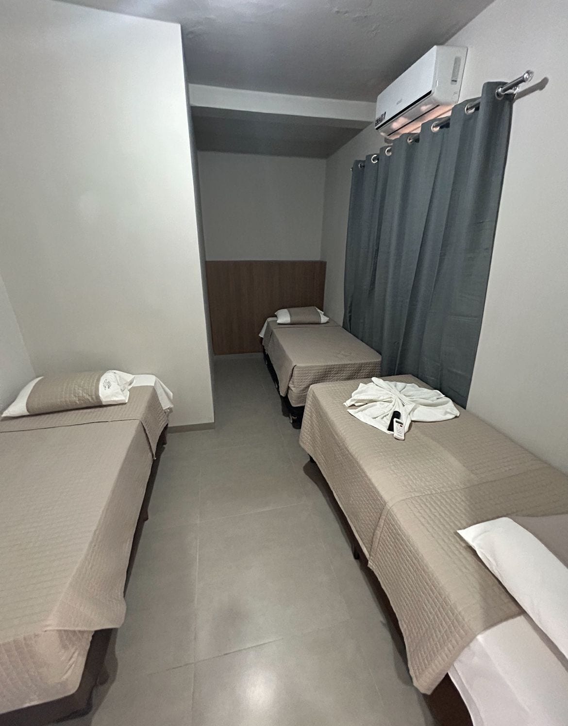 Quarto de hotel com duas camas de solteiro, uma cabeceira de madeira, cortina cinza e ar-condicionado instalado no teto.