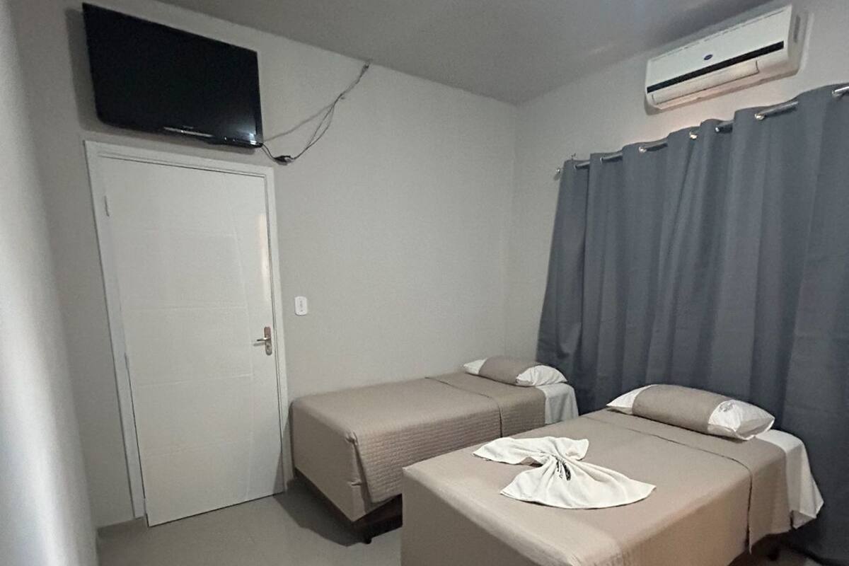 A small, clean room with two single beds, a TV mounted on the wall, and an air conditioning unit.