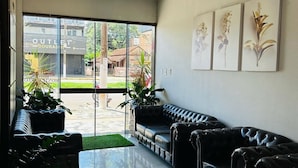 A modern interior with a black leather sofa, a glass door leading outside, and three framed floral paintings on the wall. - HOTEL MONTE CARLO (Dourados)
