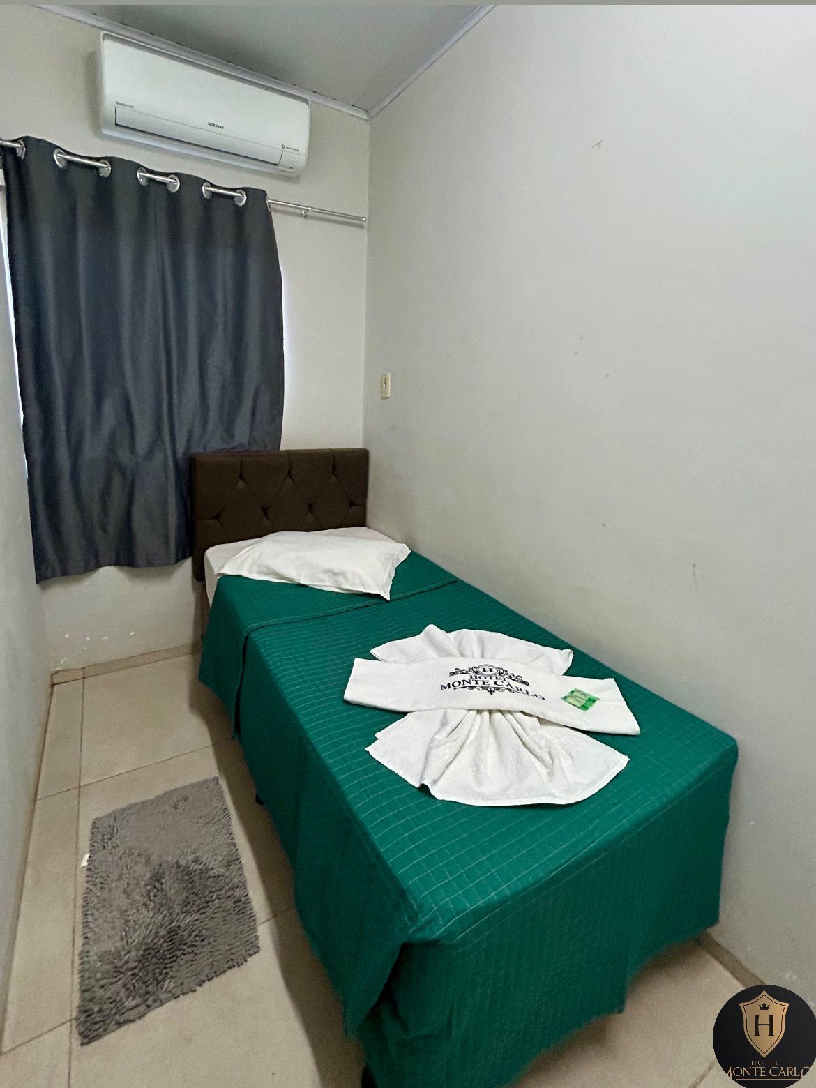 A small, single-bed room with a green bedspread, a brown headboard, and a grey rug. There is a white air conditioning unit on the wall and a grey curtain.