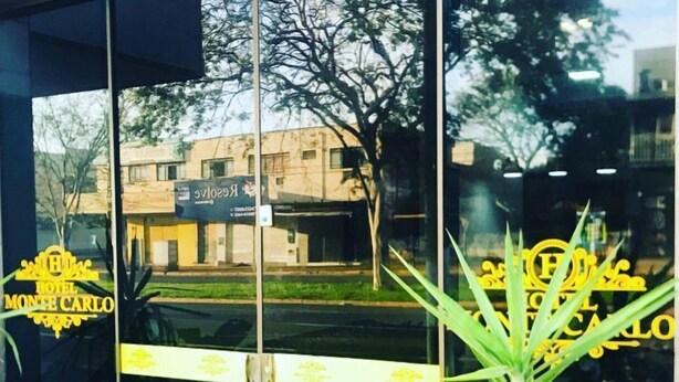 A hotel entrance with a glass door, a potted plant, and a reflection of the hotel name and a building in the background. - HOTEL MONTE CARLO (Dourados)