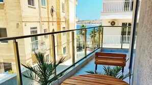 Un balcó amb taula i bancs de fusta, plantes en testos i vista al mar i edificis.