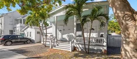 A white house with a porch, palm trees, and a car parked in front.