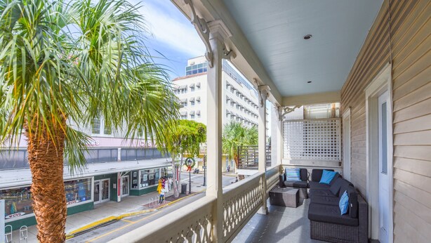 A balcony with a wicker sofa and a palm tree, overlooking a street with buildings and a CVS store. - Duval Dream Loft by Brightwild Right on Duval (Key West)