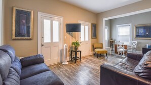 A living room with a dark leather couch, a small table with a plant, a flat-screen TV, and a dining area with a yellow chair and white table. - Duval Dream Loft by Brightwild Right on Duval (Key West)