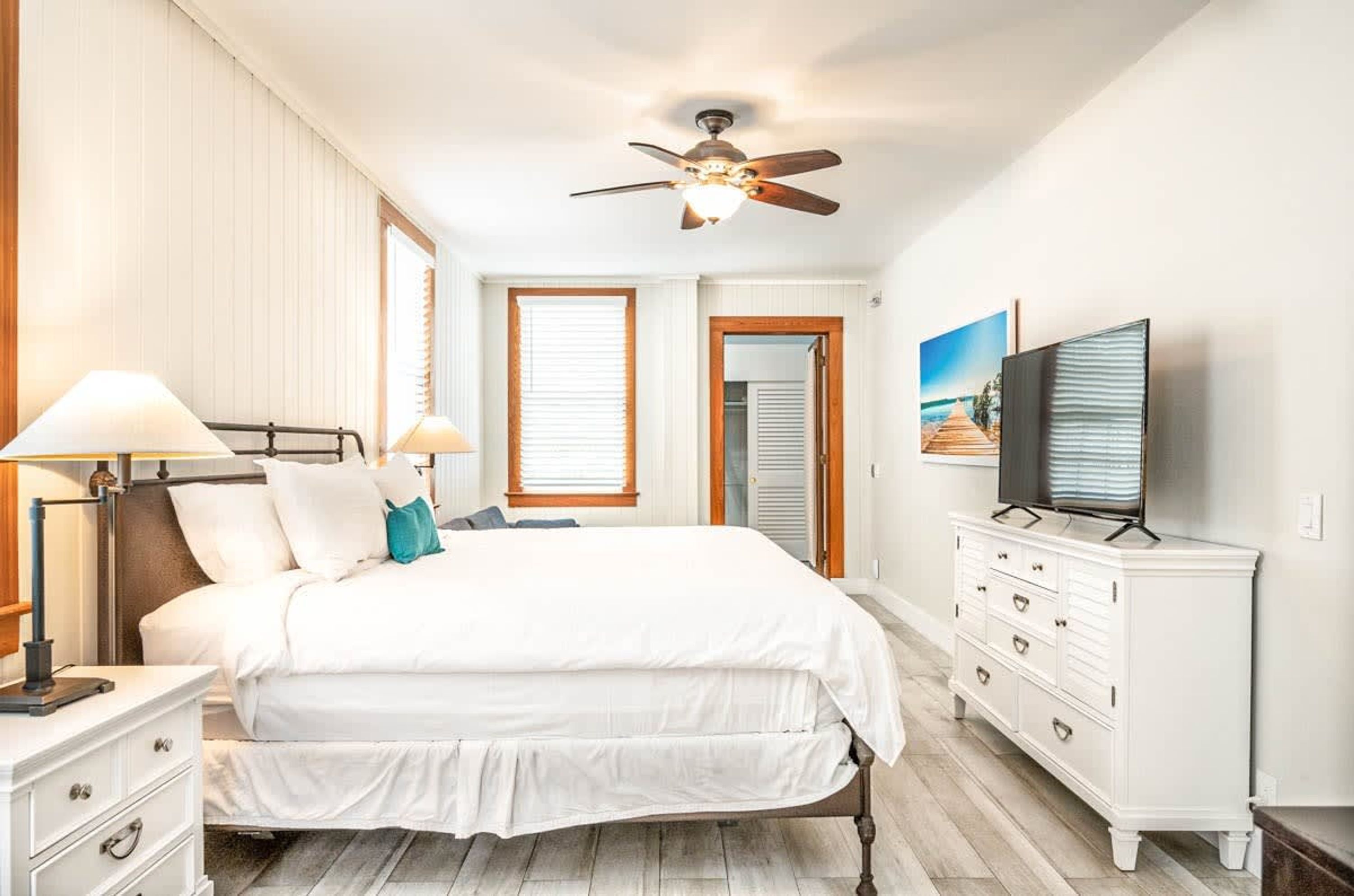 A bedroom with a bed, bedside lamps, a ceiling fan, a dresser, and a television.