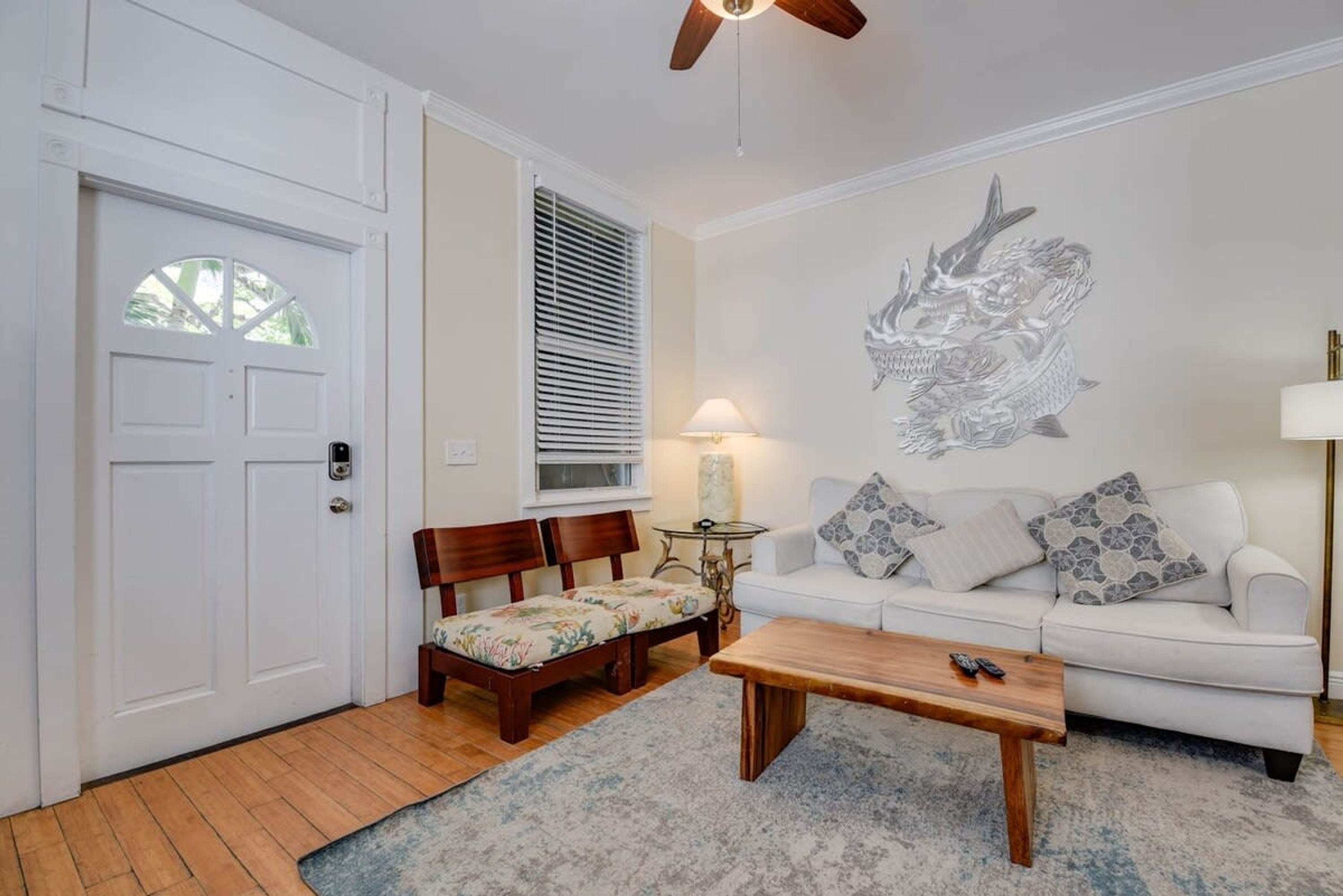 A living room with a white door, a wooden bench, a white sofa, a coffee table, and a wall mural.