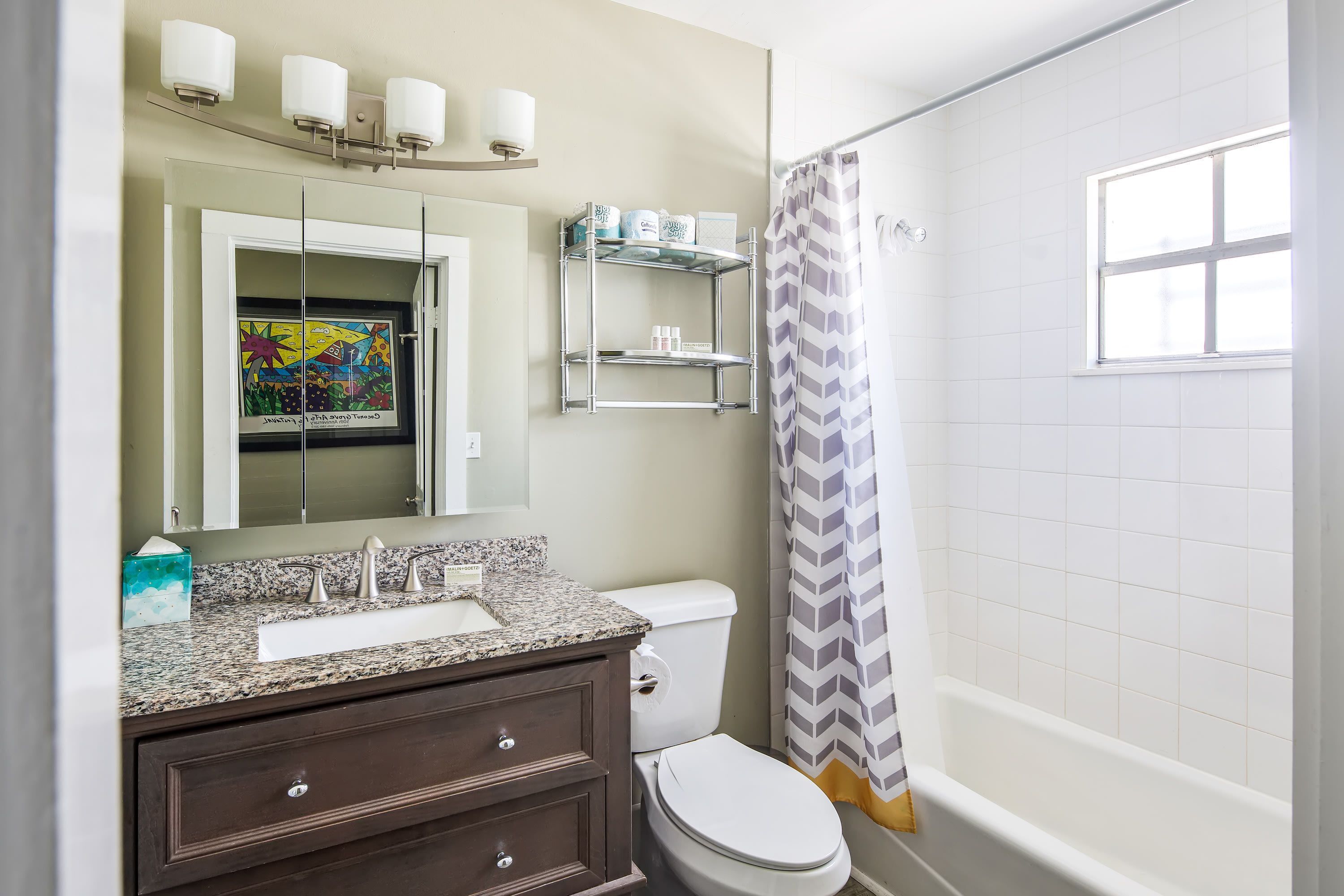 A bathroom with a white toilet, a granite countertop sink, a dark wooden vanity, a mirror with lights, a shower with a chevron curtain, and a window.