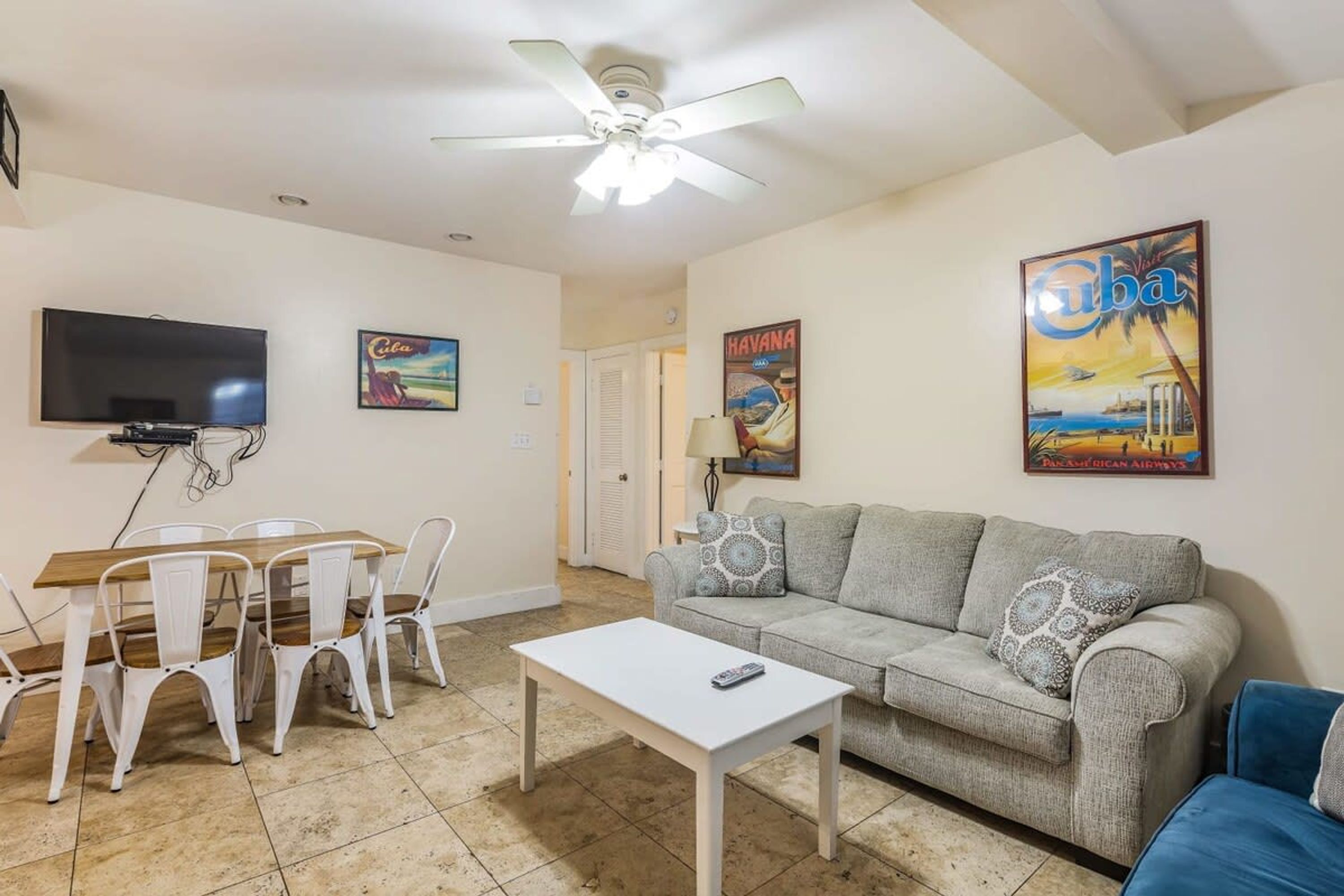 A living room with a ceiling fan, a TV, a dining table with chairs, a sofa, and a coffee table. There are posters on the walls and a lamp on a side table.