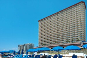 A high-rise hotel with multiple balconies, a beach with sun loungers and umbrellas, and a clear blue sky. - Coastal Condo 607 by Brightwild Beachfront (Panama City Beach)