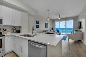 A modern kitchen with white cabinetry, a large island with a sink, and a view of the ocean through the sliding glass doors. - Coastal Condo 607 by Brightwild Beachfront (Panama City Beach)