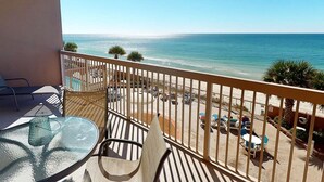 A balcony with a glass table and chairs, overlooking a beach with a clear view of the ocean and a parking area with several cars. - Coastal Condo 707 by Brightwild Beachfront (Panama City Beach)
