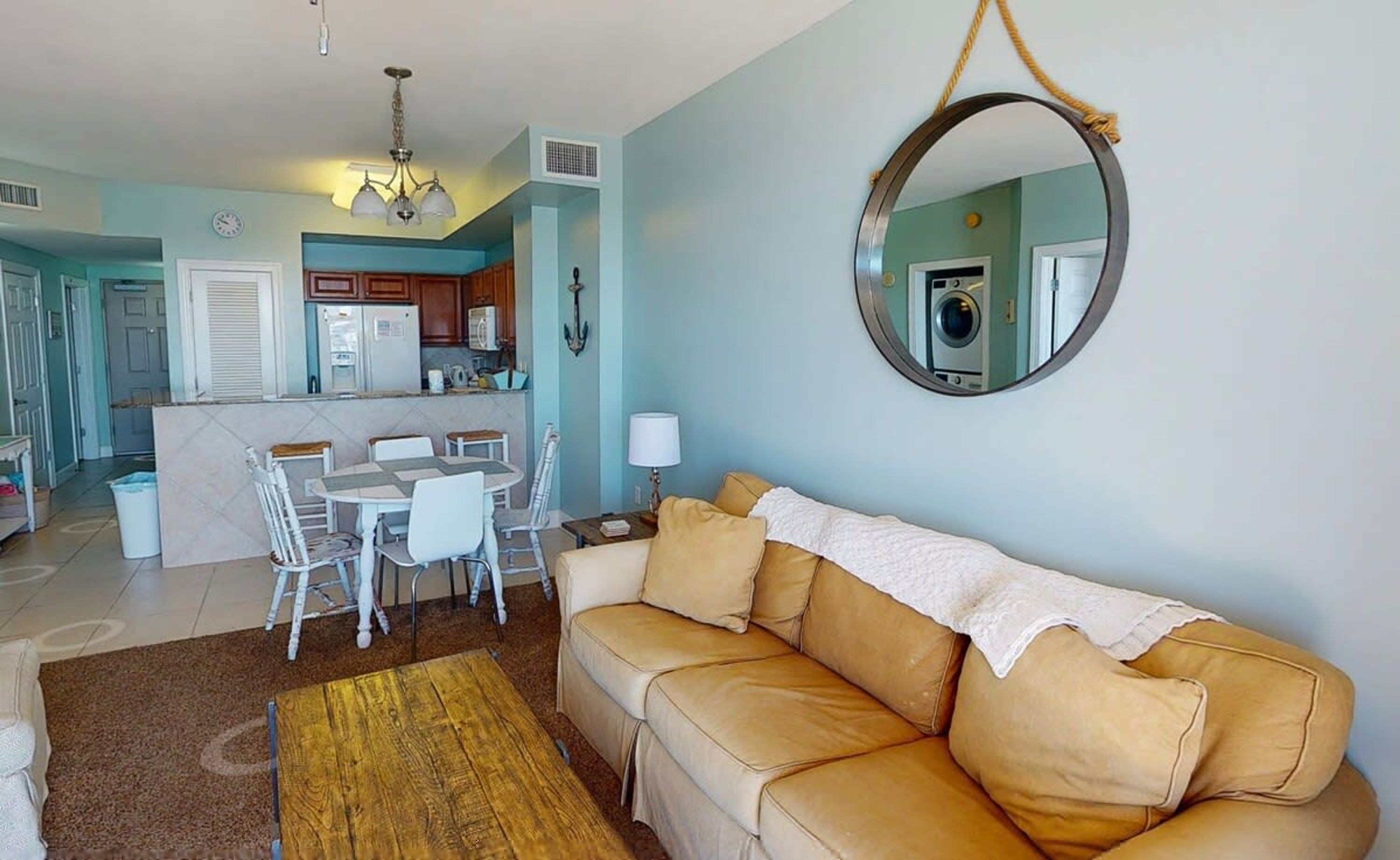 A living room with a beige sofa, a dining table with chairs, a kitchen area with cabinets and appliances, and a round mirror on the wall.
