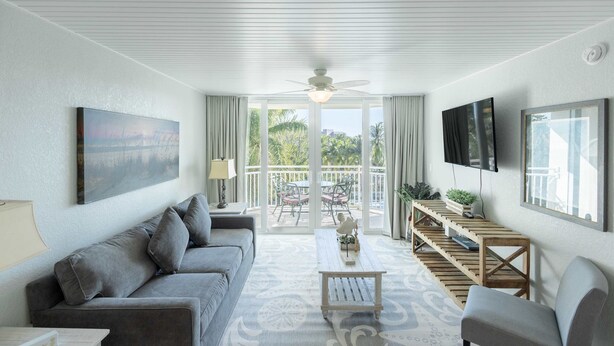 A modern living room with a grey sofa, a coffee table, and a TV mounted on the wall. There is a balcony with outdoor furniture visible through the glass doors. - The Exuma Cay by Brightwild Pool View Parking (Key West)