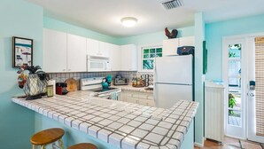 A kitchen with white cabinets, a tiled backsplash, a microwave, a refrigerator, and a window with a view of greenery. - Gulf Getaway by Brightwild Condo w Pool (Key West)