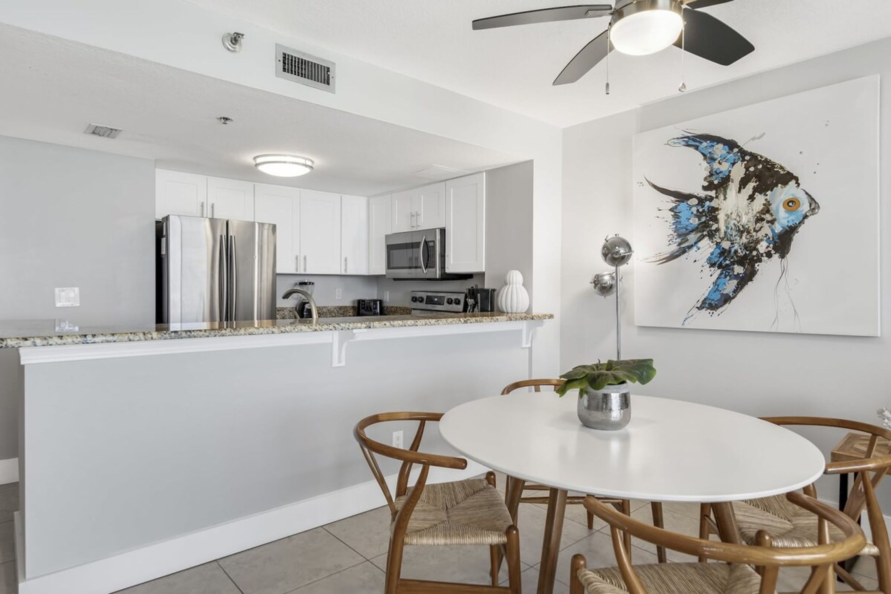 A modern kitchen with white cabinets, stainless steel appliances, a white dining table with wicker chairs, and a ceiling fan.