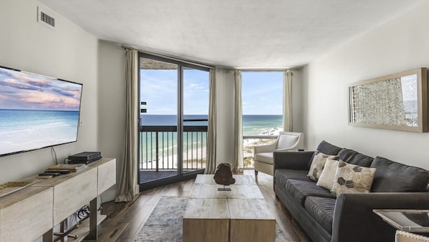 A modern living room with a large TV, a wooden coffee table, a dark sofa, and a view of the ocean through large windows.
