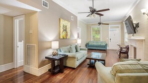 A living room with hardwood flooring, a ceiling fan, a fireplace, and a television mounted on the wall. - Bananas Foster by Brightwild Epic Guesthouse (Key West)