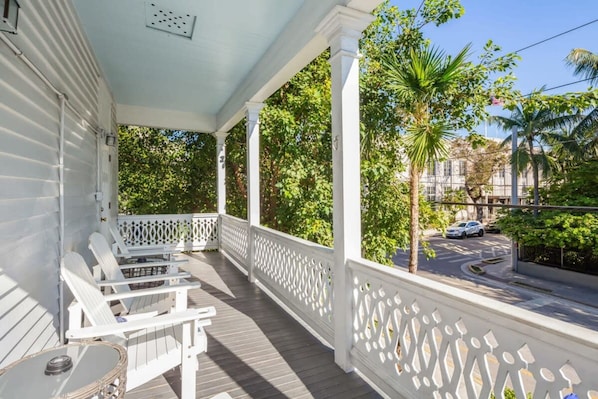 A covered porch with white railings and seating, overlooking a street with palm trees and parked cars. - Bananas Foster by Brightwild Epic Guesthouse (Key West)