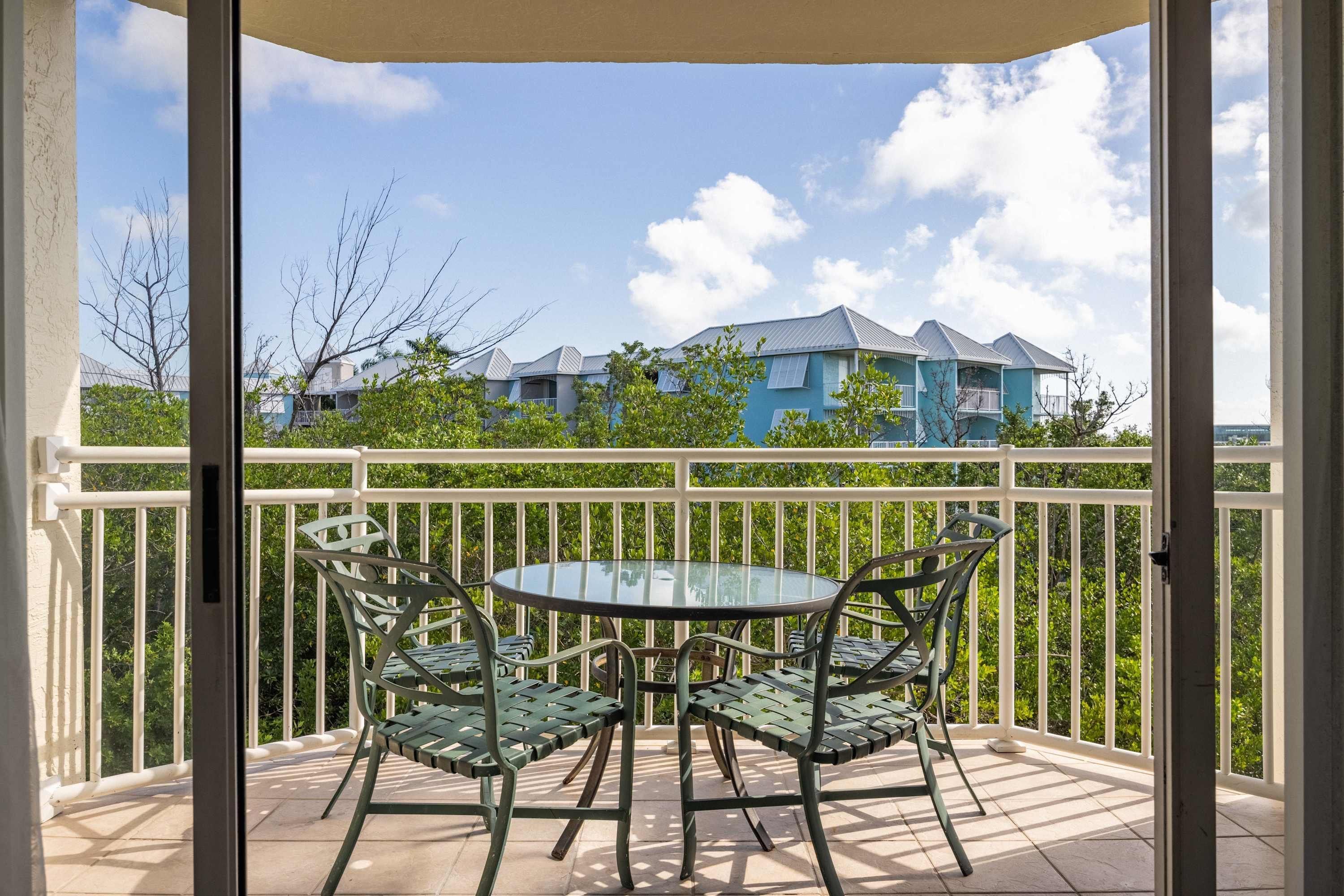 A balcony with a glass table and chairs, overlooking a residential area with blue buildings.