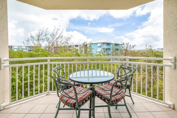 A balcony with a round table and four chairs, surrounded by a white railing, overlooking a residential area with buildings and greenery.