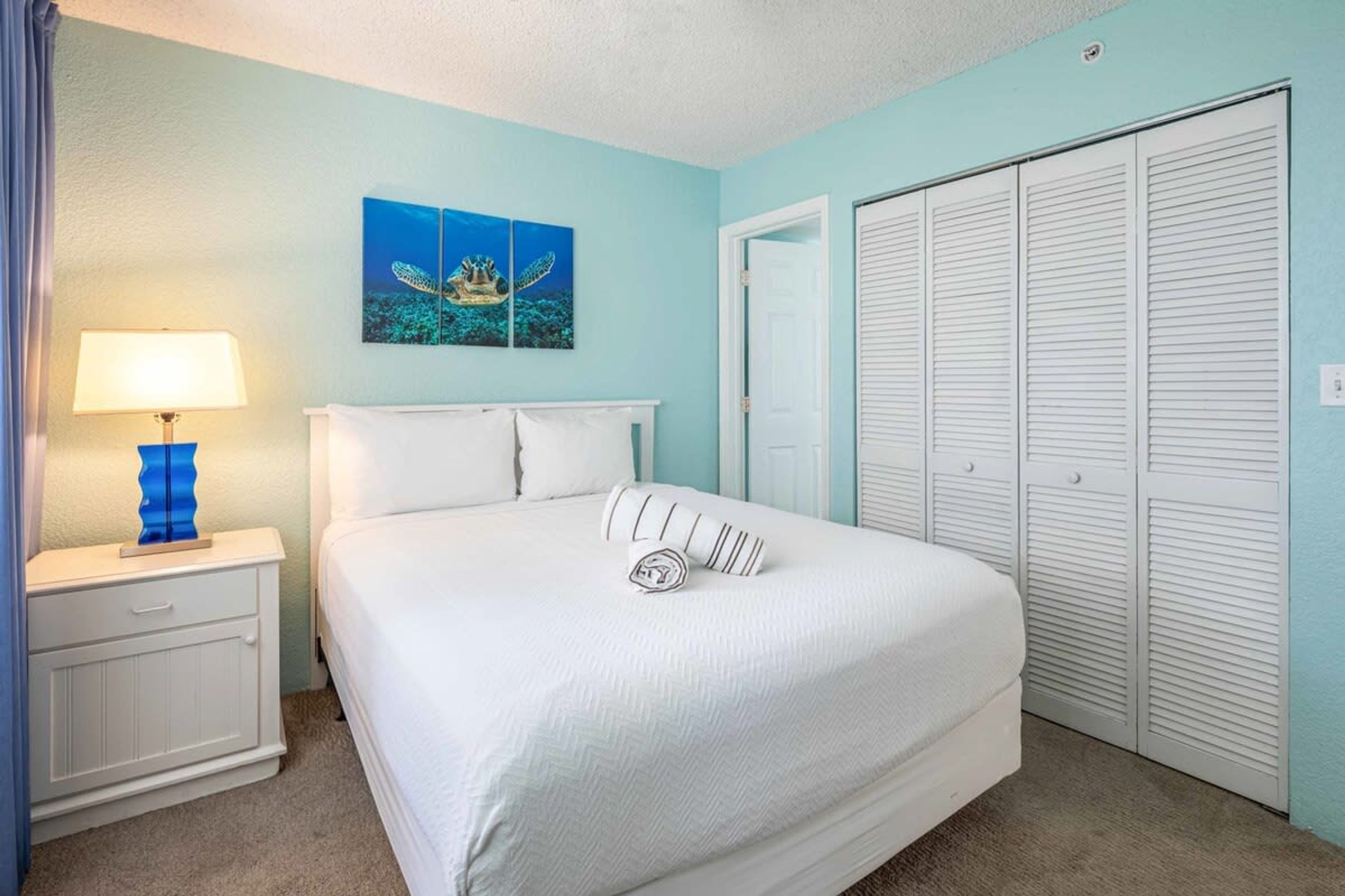 A neatly made bed with white bedding and striped pillows, a bedside lamp, a wall-mounted artwork, and a set of white louvered closets.