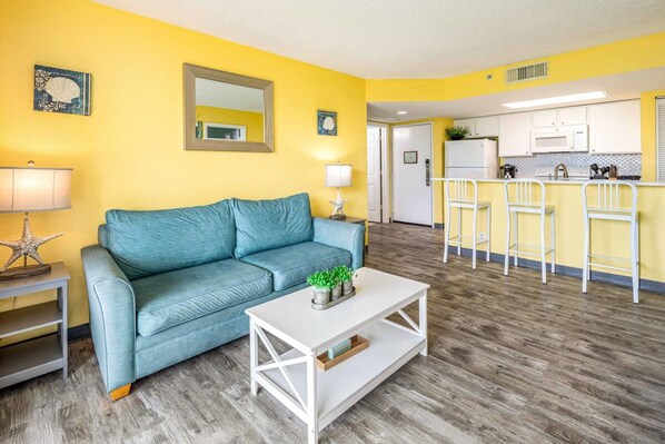 A living room with a blue sofa, a white coffee table, and a kitchen area with a bar counter and white bar stools. - Tortuga Suite Sparkling Pool Hot Tub (Key West)