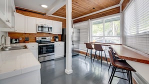 A modern kitchen with white cabinets, stainless steel appliances, and a wooden ceiling. There is a bar area with stools and a window with blinds. - Commodore s Compound by Brightwild Huge Home (Key West)