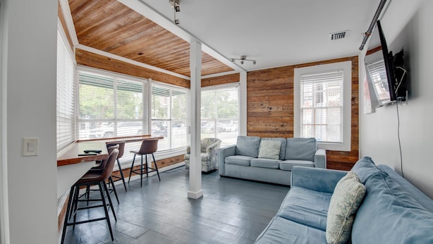 A modern living room with a wooden ceiling, a grey sofa, a dining area with chairs, and large windows with blinds. - Commodore s Compound by Brightwild Huge Home (Key West)