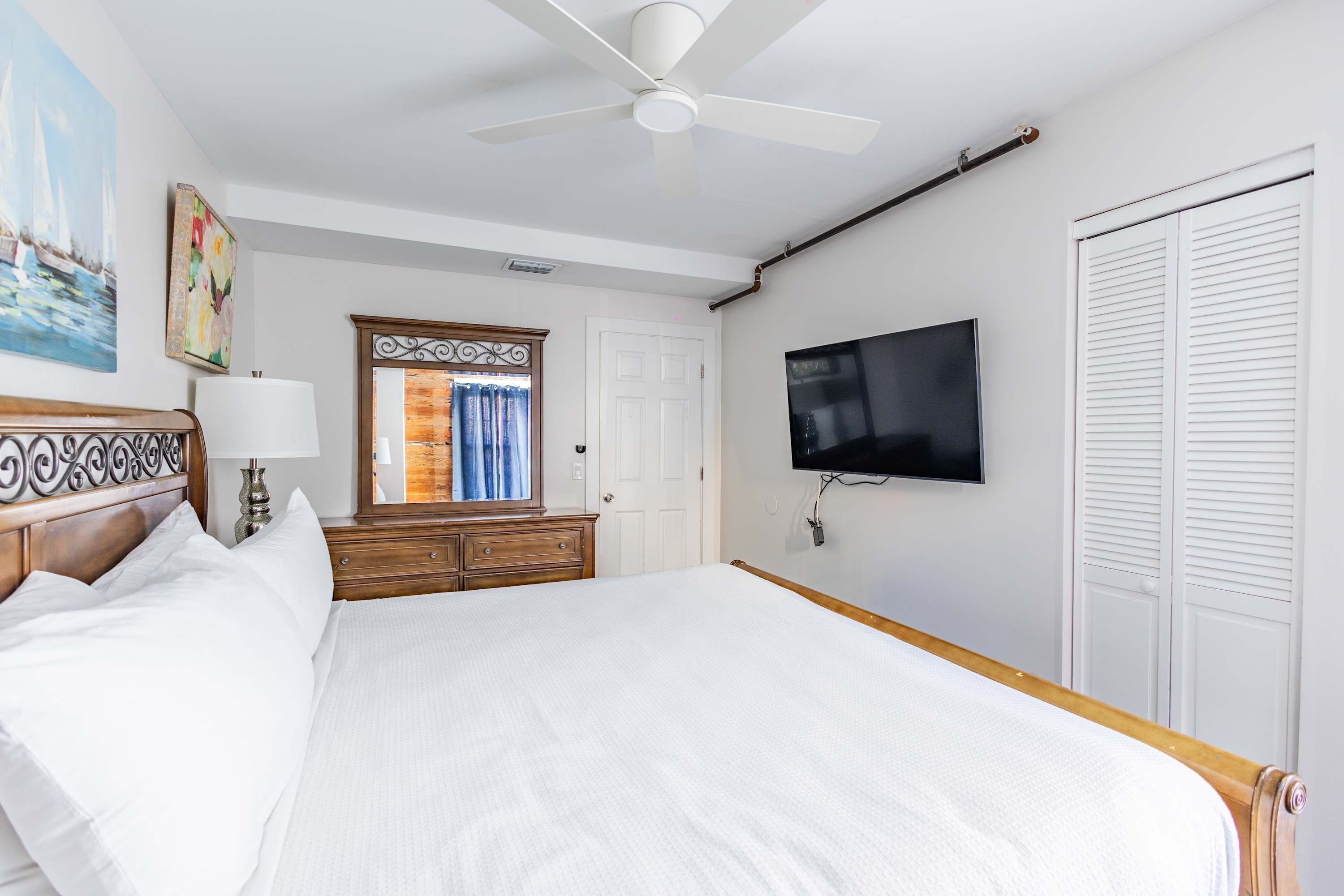 A hotel room with a bed, a ceiling fan, a TV, a dresser, a mirror, and a door.