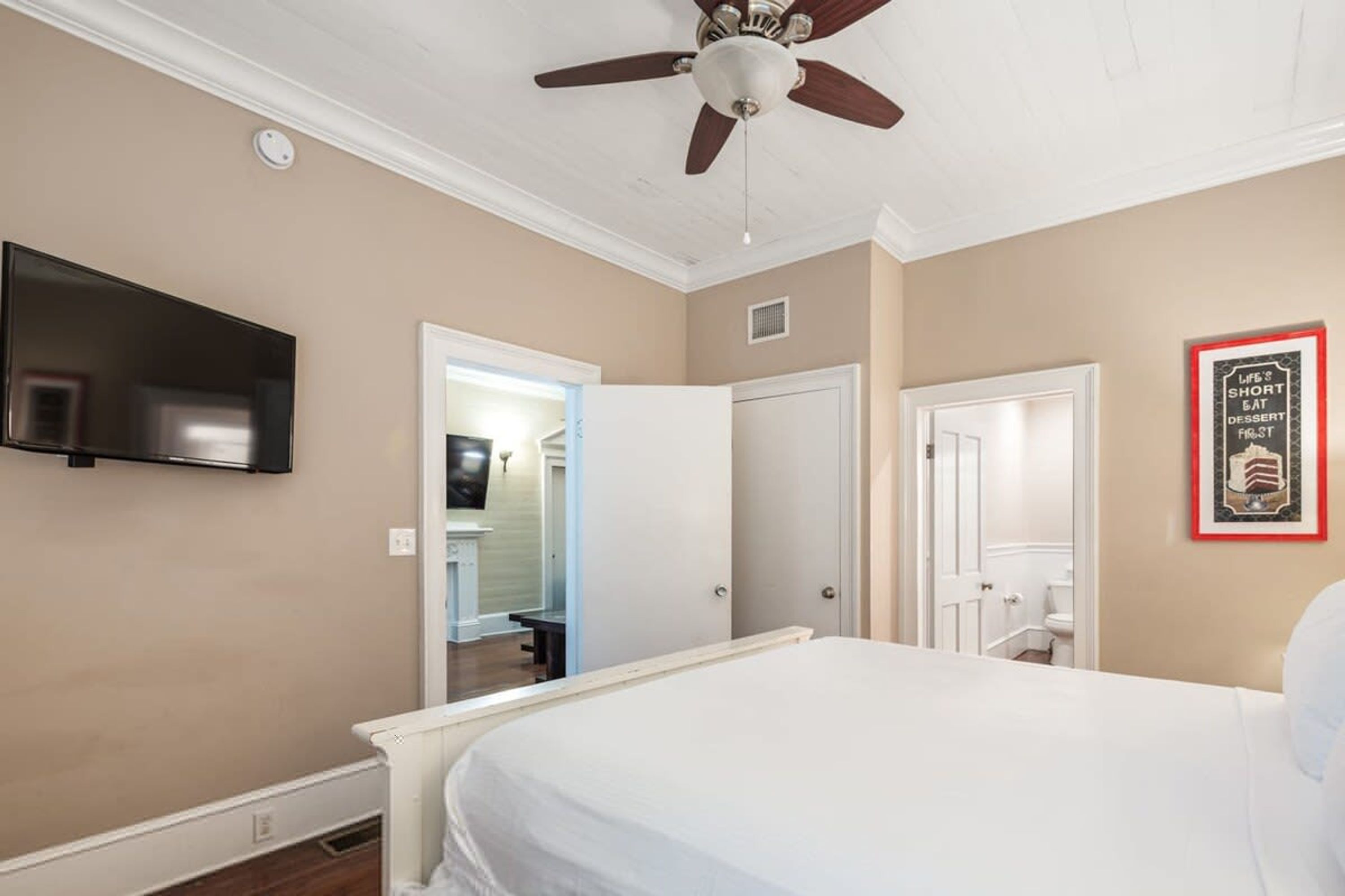 A bedroom with a bed, a ceiling fan, a TV mounted on the wall, and a door leading to a bathroom.