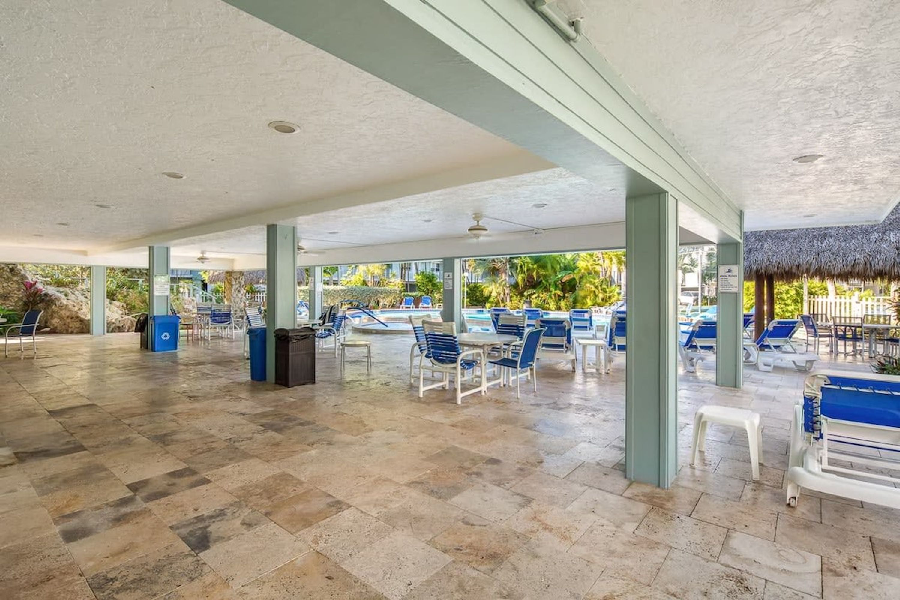 An outdoor pool area with blue and white lounge chairs, a covered seating area, and a thatched-roof structure.